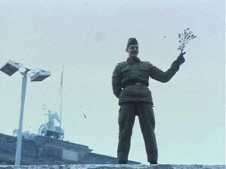 East German guard on top of Wall waves with a bouquet of flowers